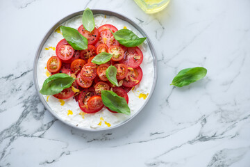 Plate with stracciatella di bufala, fresh tomatoes and green basil, above view on a white marble background, horizontal shot