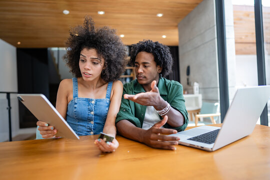 A young couple of African descent sit at a table in their modern apartment, looking stressed and discussing financial issues. Both are using electronic devices, visibly concerned about their situation