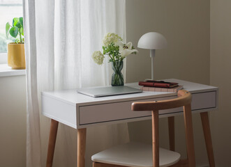 A white table with a laptop, a lamp, notebooks, a bouquet of flowers by the window in the living room. A cozy workplace in a minimalist style
