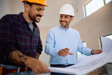 Happy housing project manager examining blueprints with  worker at construction site.