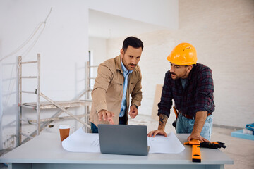Mid adult man and building contractor examining housing plans on laptop at construction site.