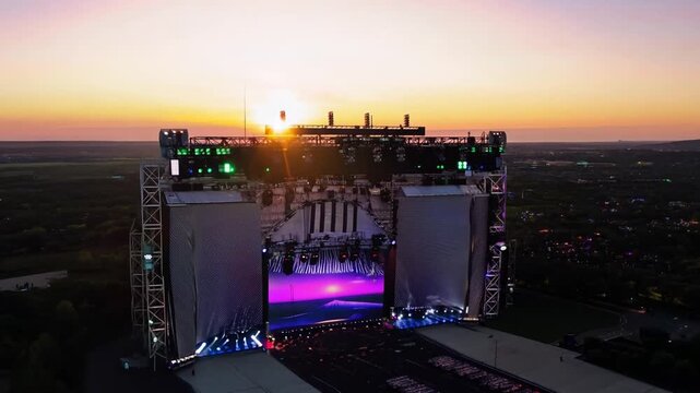 An aerial view of a large outdoor stage set up for a concert or event, with a giant screen displaying a sunset scene. The stage is surrounded by a large crowd.