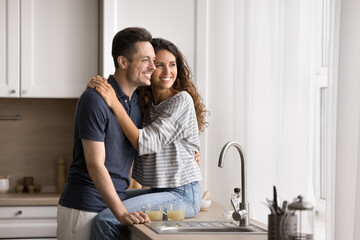 Harmonic millennial Hispanic couple in love spend time together in modern kitchen, hugging, smiling, looking into distance, daydreaming about bright joint future at new house. Marriage, relationships