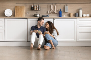 Young Hispanic couple resting seated on warm floor in fashionable kitchen, enjoy pleasant...
