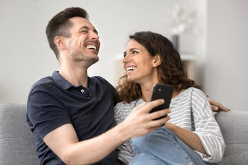 Obraz premium Cheerful young wife showing funny videos on social media network on cellphone to laughing husband, sit together on sofa, resting on weekend using new mobile app, giggling, having fun. Tech, leisure