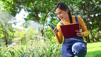Beautiful young asian woman is sitting studying natural plants and herbs garden using her phone take pictures flowers and herbs and writing the names plants on a clipboard with interest.