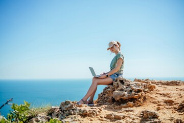 A woman is sitting on a rock by the ocean, using her laptop