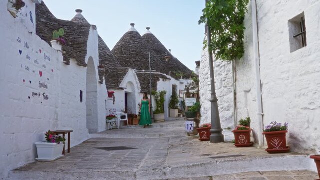 A young hispanic woman in a green dress walks down a picturesque street lined with trulli houses in the old town of alberobello, puglia, italy, on a sunny day.