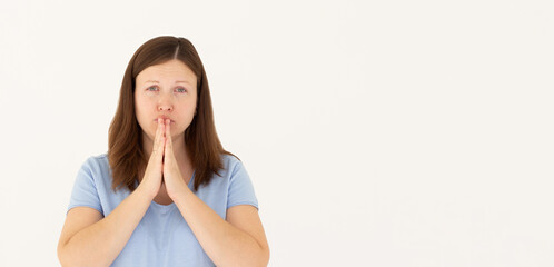 Portrait of imploring girl tilts head, purses lips, looks sadly at camera, makes praying gesture,...