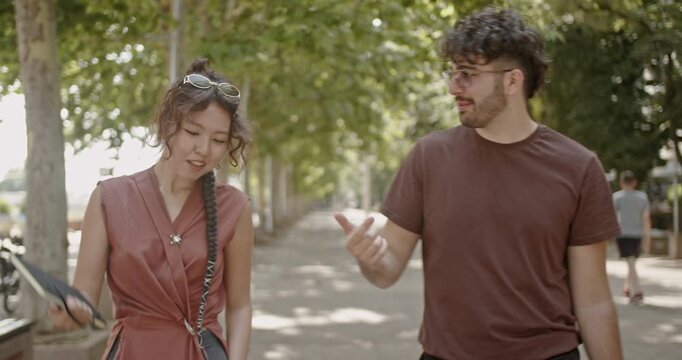 Two young business friends, a man and a woman, enjoying a casual conversation while walking together in a beautiful, sunny, tree-lined park.