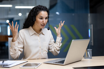 Frustrated businesswoman wearing headset gesturing at laptop during virtual meeting in modern...
