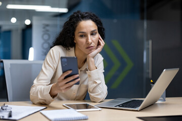 Bored businesswoman sitting at desk, staring at smartphone. Office environment with laptop, tablet,...