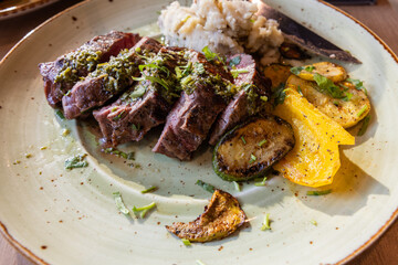 A plate of meat with a side of mashed potatoes and vegetables. The meat is cooked and seasoned perfectly, and the vegetables are roasted.