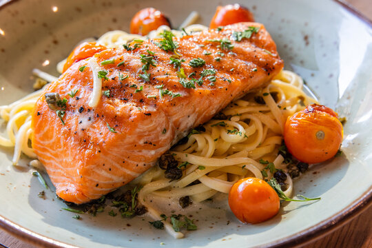 A plate of pasta with a piece of salmon on top. The salmon is seasoned with herbs and spices. The pasta is cooked and served in a bowl. The dish looks delicious and appetizing