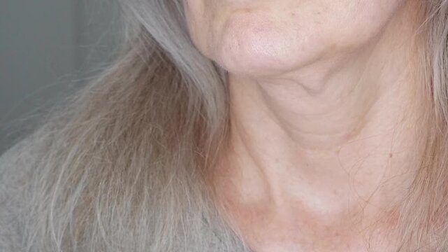 Close-up of a woman's neck with gray hair. The woman touches her loose and saggy skin on her chin and neck. The concept of cosmetology and plastic surgery. The concept of skin care in middle age.