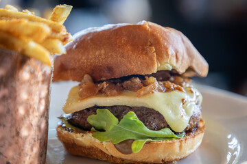 Beef tenderloin burger with lettuce and cheese sits on a white plate next to a metal bowl with fries. The burger is the main focus of the image, and the bowl is a decorative element that adds a touch 