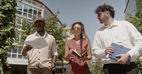 A diverse team of business colleagues is walking together outdoors, engaging in a lively discussion as they enjoy a sunny day in the city, surrounded by the vibrant urban landscape.