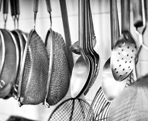Black and white image of commercial kitchen utensils including spoons and strainers hanging on a...