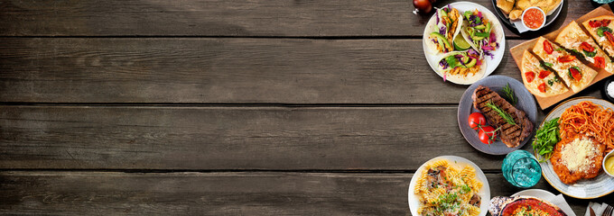 Side border of various delicious foods. Overhead view on a dark wood banner background. Copy space. Pastas, flatbreads, tacos, steak and restaurant theme dishes.