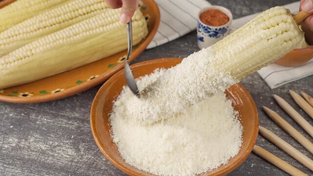 Mexican woman putting grated aged cheese on top of Mexican corn prepared with mayonnaise
