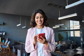 Young Indian woman manager using cell phone mobile AI app for call, business communication, trading. Focused Latin Hispanic female businesswoman holding smartphone for online work, standing in office