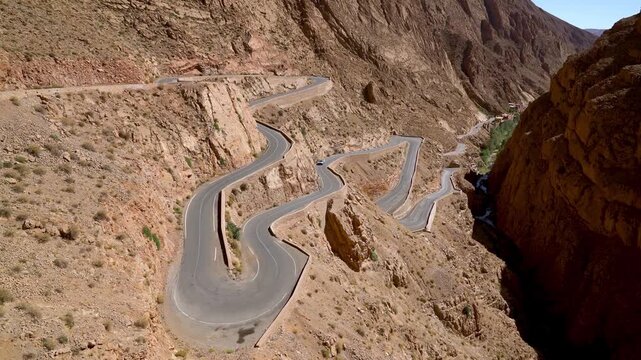 Unidentified car driving through winding road in Dades Gorges, a series of canyons formed by the Dades River. Red and orange rock formations create a dramatic and picturesque landscape.