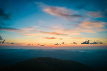 Fototapeta premium Sun sets behind rolling hills, casting warm, golden glow across sky. Wispy clouds, tinged with hues of orange and yellow, stretch across deepening blue sky. Serene and expansive landscape.