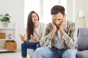 A young man sitting on the couch covers his ears from a womans scream. The wife scolds her husband,...