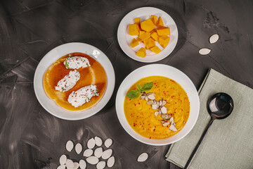 Traditional vegetable cream pumpkin and carrot soup in a white bowl, sliced pumpkin slices on a plate and pancake curd quenelles on a gray graphite background with pumpkin seeds
