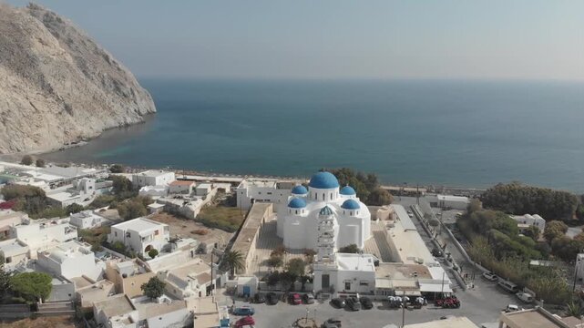 Aerial panorama drone view of Church of the Holy Cross, near Perissa black pebble beach near mountain, white houses with hotels and restaurants. Santorini Island, Greece. Aegean Sea, Mediterranean.
