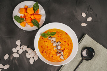Vegetarian dish: traditional vegetable cream soup of pumpkin and carrot with pumpkin seeds and mint in a white bowl and sliced pumpkin on a white plate on a gray background with a green napkin