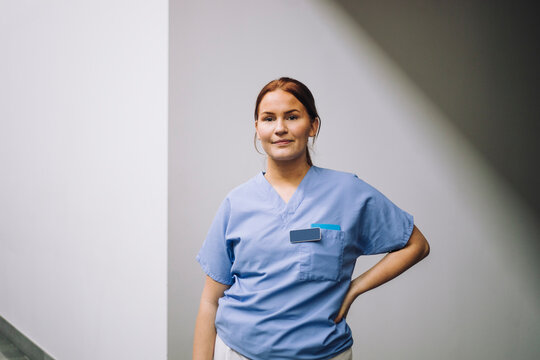 Portrait of smiling young female medical trainee standing with hand on hip near wall in hospital
