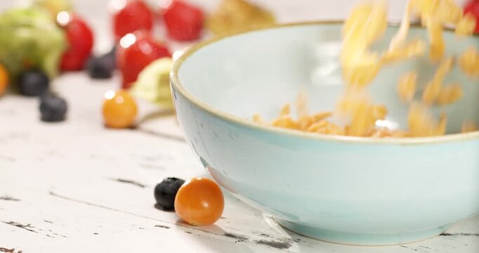 Pouring corn flakes into a bowl against the background of various berries lying on the breakfast table.