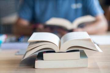 Closeup image with bokeh of an open book standing on other books on a desk and a man out of focus in the background reading another book in a library