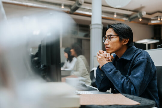 Focused male entrepreneur with hands clasped looking at computer screen while sitting in office