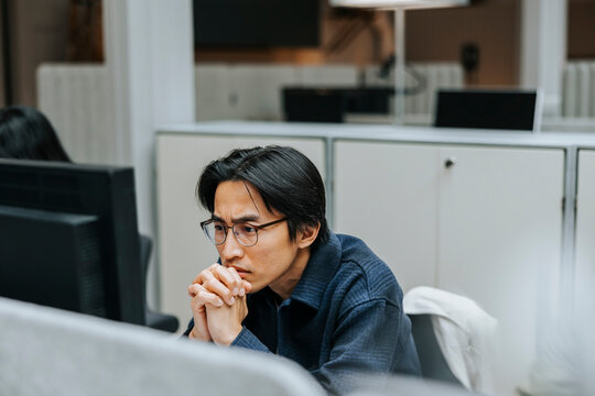 Focused male business professional with hands clasped looking at computer screen in office