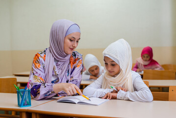 Female hijab Muslim teacher helps school kid to finish the lesson during the class in the classroom. 