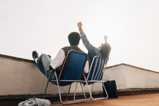 Low angle view of man cheering while sitting with male friend on chair against sky