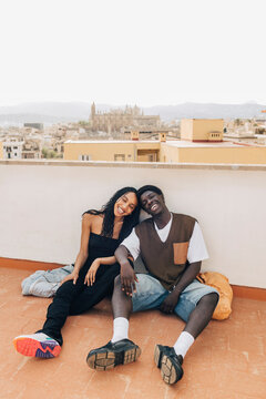Carefree male and female friends sitting on floor near retaining wall on rooftop