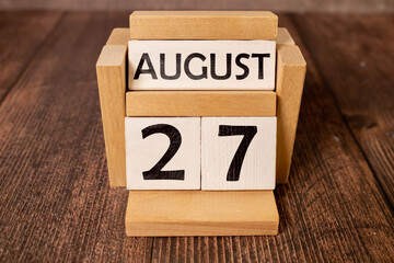 Wooden block calendar with the date August 27 and spikelets of wheat on a beige concrete background.