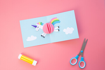 Handcrafted three-dimensional paper model of a unicorn featuring a pink body, white head with purple details like eyes and ears. Rainbow-colored mane and tail, leaping over white cloud shapes