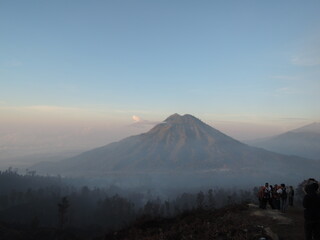 Panoramic landscape view of Mount Bromo volcano in Indonesia, surrounded by smoke and fog. The iconic volcano stands tall amidst a dramatic setting, with misty fogs enhancing the mystical atmosphere.
