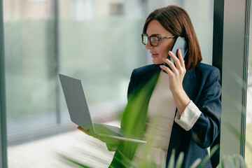 A businesswoman is multitasking using her phone and laptop in a contemporary office setting