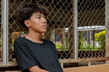 Asian teenboy in a black shirt sits with his knees pressed against a metal fence panel in a...
