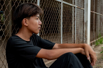 Asian teenboy in a black shirt sits with his knees pressed against a metal fence panel in a...