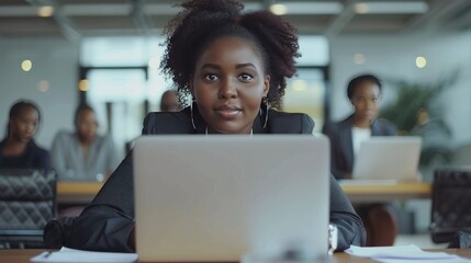 Attractive african young confident businesswoman sitting at the office table with group of colleagues in the background, working on laptop computer - generative ai