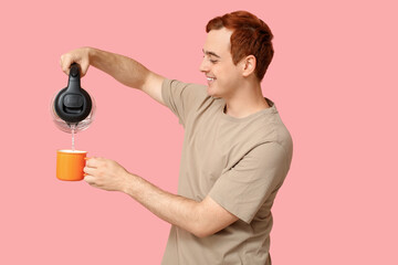 Handsome young man pouring hot water from modern electric kettle into mug on pink background