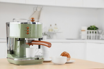 Modern green coffee machine with bowl of sugar, cups and cupcake on table in kitchen