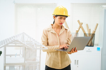 An Asian female architect analyzes blueprints at her desk in the office. She specializes in house design, focusing on roofs and walls, and collaborates with construction contractors in her company.