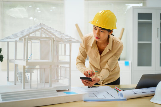 An Asian female architect analyzes blueprints at her desk in the office. She specializes in house design, focusing on roofs and walls, and collaborates with construction contractors in her company.